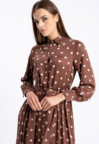 Brązowa sukienka koszulowa z długimi rękawami w białe kropki, z kołnierzykiem, wiązaniem w talii i zapięciem na guziki, o gładkiej fakturze materiału.