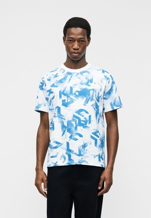 Νέος άνδρας που φοράει λευκό κοντομάνικο μπλουζάκι με μπλε αφηρημένα γεωμετρικά σχέδια, στέκεται μπροστά σε ένα απλό ανοιχτό γκρι φόντο.