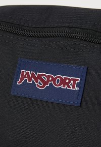 Zaino in tessuto nero con superficie testurizzata, dotato di un'etichetta rettangolare blu navy che mostra il logo "JANSPORT" in lettere bianche e rosse.
