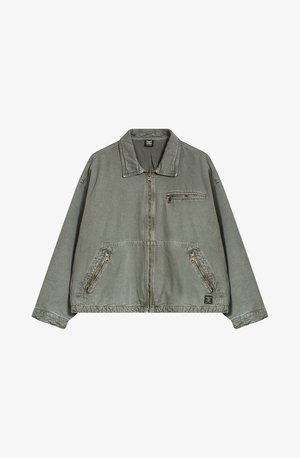 Olajzöld dzseki, gallérral, elülső cipzárral és két oldalsó zsebbel. Tartalmaz egy cipzáros mellzsebet és kontrasztos varrási részleteket.