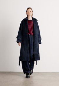 Cappotto lungo in misto lana blu marino con larghe rever, apertura frontale e maniche lunghe, indossato sopra una camicia a righe rossa e blu marino e pantaloni scuri larghi.