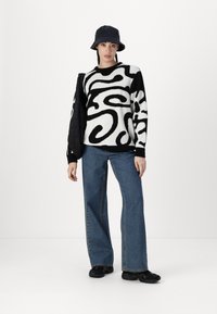 Pull en tricot à motifs abstraits noir et blanc, associé à un jean large bleu foncé et des baskets noires, complété par un chapeau seau noir.