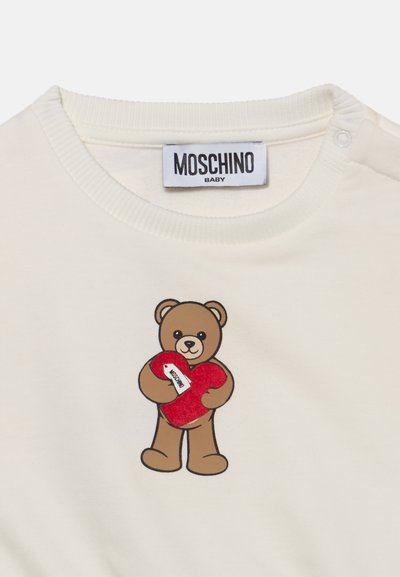 Ρούχο μωρού σε εκρού χρώμα με ετικέτα Moschino και μια αρκουδίτσα που κρατά μια κόκκινη καρδιά με μικρή ετικέτα Moschino πάνω της.