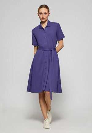 Robe courte violette à manches courtes avec col, front boutonné, ceinture nouée à la taille et jupe évasée. Portée avec des baskets blanches.