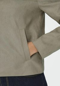 Ljusgrå falsk mockajacka med minimalistisk design, som har en sidficka och en slät yta. Detaljerat broderi längs sömmarna.