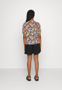 Blusa com padrão floral e mangas curtas bufantes em cores vibrantes sobre um fundo azul marinho; combinada com calções pretos e sandálias planas.