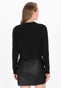 Schwarzer, verkürzter Pullover mit langen Ärmeln, strukturiertem Stoff und silbernen Ösen-Schnürdetails an den Schultern. Kombiniert mit einem schwarzen Rock aus Kunstleder.