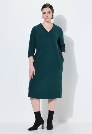 Abito verde teal con scollatura a V, maniche a tre quarti e tasche laterali. La modella indossa calzini neri alla caviglia con scarpe a tacco largo.