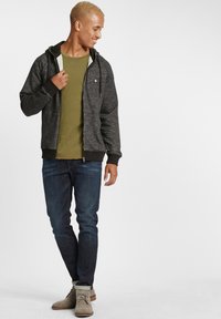 Sivi zip-up dukserica s crnim detaljima, maslinasto zeleni majica ispod, plave traperice i bež cipele. Tekstuirana tkanina, ležeran kroj.