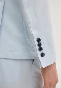 Manche de costume bleu clair avec quatre boutons foncés près du poignet, montrant une partie du pantalon assorti et la main d'une personne sur le côté.