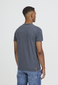 Camiseta gris de manga corta con cuello redondo, que presenta una textura sutil. Una pequeña etiqueta de la marca en marrón es visible cerca del dobladillo inferior.
