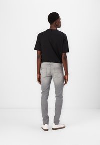 Black cotton t-shirt, short sleeves, paired with light gray tapered jeans. Model wears white sneakers, viewed from behind against a white background.