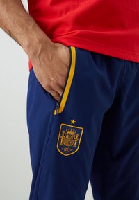 Morske atletske hlače s žutim akcentima i džepovima na zatvarač, s žutim logotipom španjolske nogometne reprezentacije uz džep.