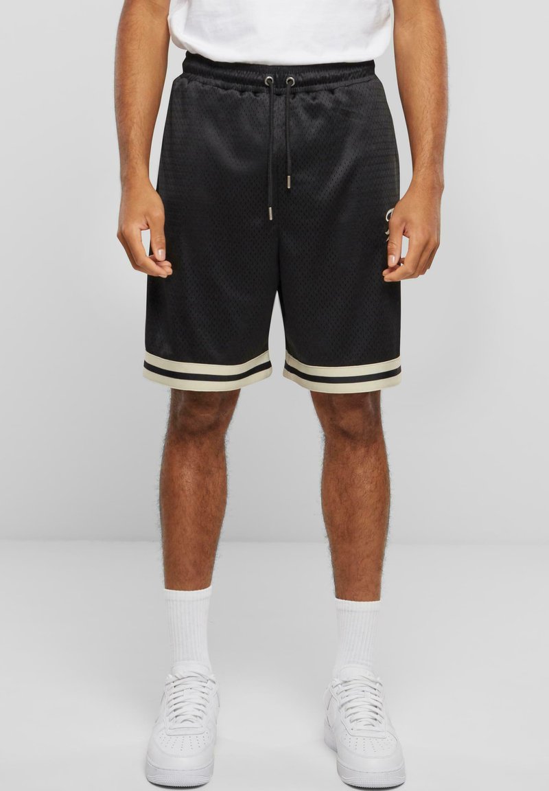 Svart idrettsshorts med mesh-struktur, hvite striper ved kanten og snøring i livet. Stilfullt kombinert med hvite joggesko og sokker.