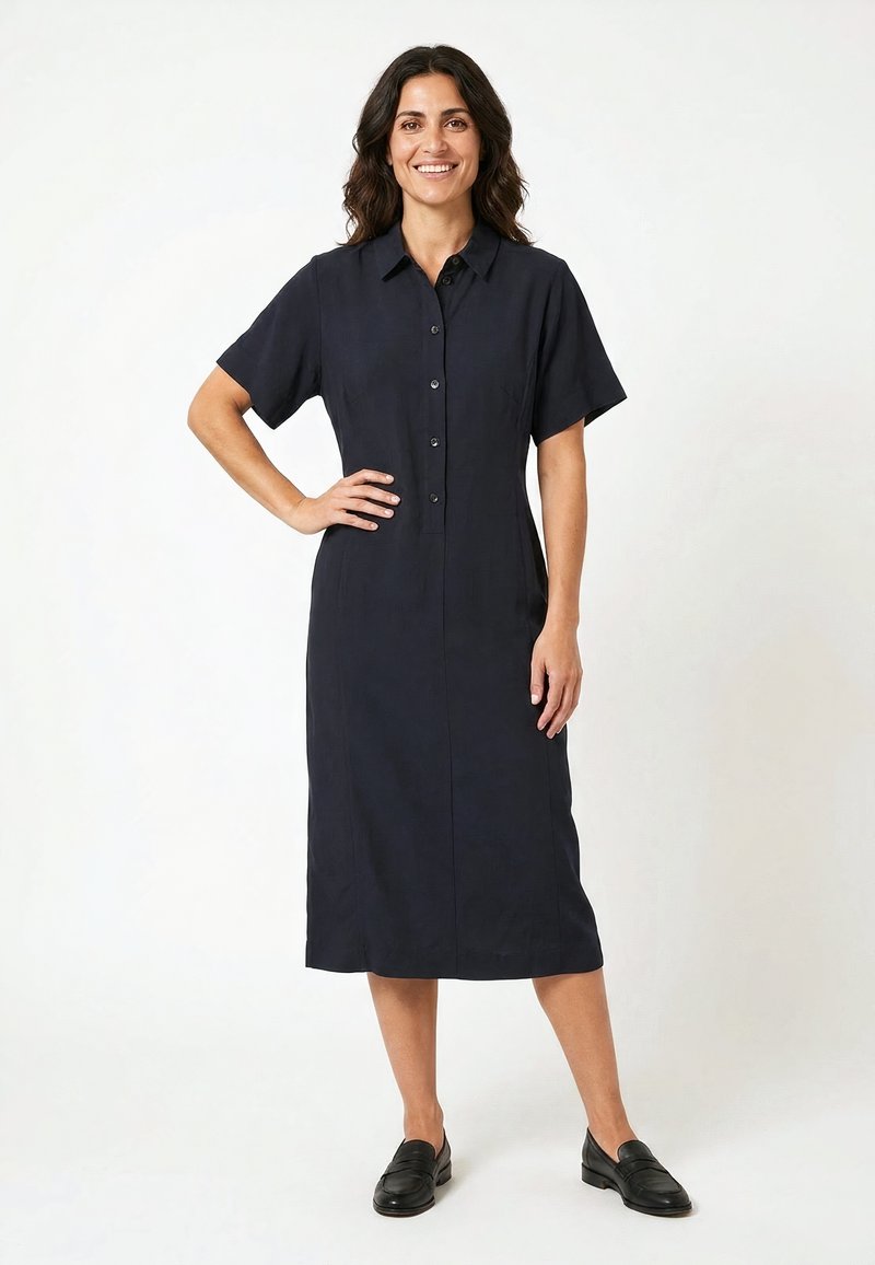Femme aux cheveux foncés et ondulés, vêtue d'une robe noire boutonnée à manches courtes et longueur genou, ainsi que des chaussures noires faciles à enfiler, souriante avec une main sur la hanche.