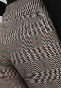 Szare spodnie w kratę z pomarańczowymi i czarnymi akcentami. Posiadają plecioną teksturę i jedną tylną kieszeń. Brak widocznych zapięć lub dodatkowych detali.