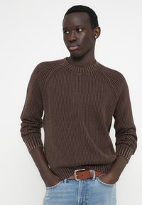 Καφέ πλεκτό πουλόβερ με ριμπ ύφανση, μανίκια ραγκλάν, στρογγυλή λαιμόκοψη και υφή με ανάγλυφη επιφάνεια, σε συνδυασμό με τζιν ανοιχτού χρώματος και καφέ ζώνη.