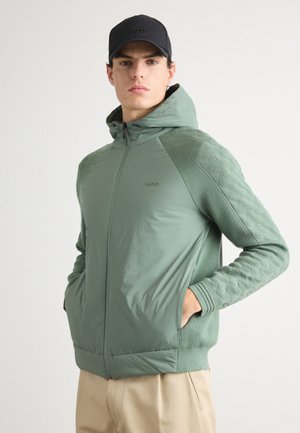 Svetlozelena zip-up kapuca s teksturiranim rukavima i žepovima, napravljena od mješavine tkanina. Ima kapuljaču i suptilan brend na grudima.