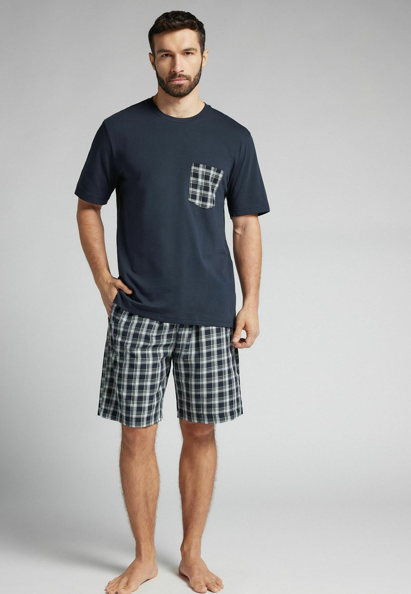 Camisa de manga corta azul marino con un bolsillo a cuadros, combinada con shorts de patrón a cuadros en azul marino, verde y blanco. Material suave de algodón.