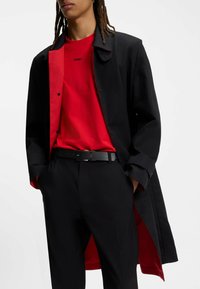 Cappotto nero con interno rosso, indossato sopra una maglietta rossa con il logo "HUGO". La cintura nera contrasta con i pantaloni scuri, evidenziando i diversi strati.