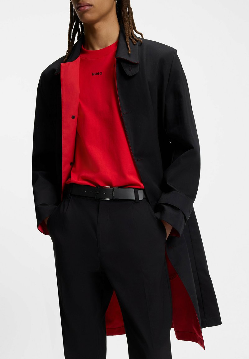Cappotto nero con interno rosso, indossato sopra una maglietta rossa con il logo "HUGO". La cintura nera contrasta con i pantaloni scuri, evidenziando i diversi strati.