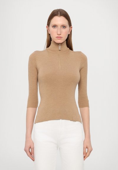 Haut en tricot beige à col roulé avec une fermeture éclair half-zip, des manches courtes, une texture côtelée et une silhouette ajustée. Un pantalon blanc est porté en dessous.