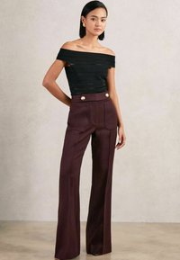 Fekete váll nélküli blúzban és magas derekú, széles szárú bordó nadrágban, arany gombokkal, áll egy sima világos háttér előtt.