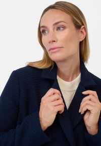 Marinblå texturerad kappa med snedkrage, bärs över en krämfärgad tröja. Motivet håller i kappans slag, med subtila sömnadsdetaljer.