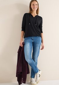 Sweat à capuche noir et léger avec un col en V et des cordons de serrage, associé à un jean bleu clair. Tenant une veste polaire bordeaux foncé ; portant des baskets blanches.