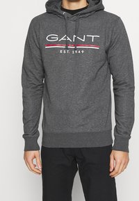 Uomo che indossa un felpa GANT grigio scuro con logo bianco e rosso e pantaloni neri, in piedi contro uno sfondo chiaro e semplice.