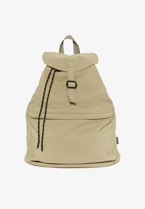 Beiger Stoff-Rucksack mit vorderer Reißverschlusstasche, schwarzem Schnallenverschluss, schwarzen Kordelzügen und Tragegriff oben.