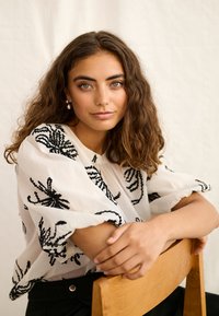 Jeune femme aux cheveux bruns bouclés portant un chemisier blanc avec une broderie noire, assise les bras reposant sur le dossier d'une chaise en bois.