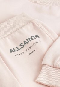 Ljusrosa sweatshirt med ribbade muddar och krage. Frontrycket inkluderar "ALLSAINTS" och text på flera språk. Mjuk textur.