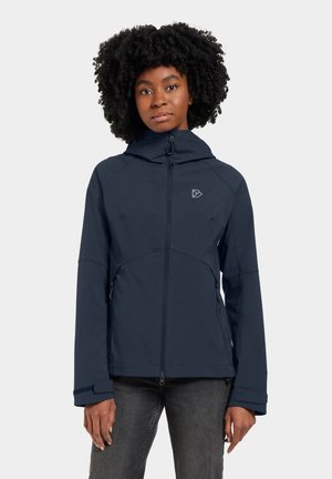 Donna con capelli ricci che indossa una giacca blu scuro con cappuccio, cerniera e tasche laterali, in piedi davanti a uno sfondo chiaro e uniforme.