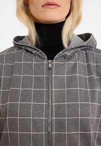 Haut à capuche gris zippé avec un motif en quadrillage blanc, doté d'une capuche et d'une fermeture éclair en argent. Porté sur un col roulé noir. Tissu lisse.