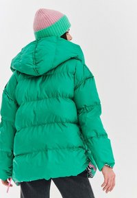 Osoba v jasne zelenej bunda s kapucňou a ružovo-zelenou pletenou čiapkou, otočená chrbtom, v čiernych nohaviciach na jednoduchom pozadí.