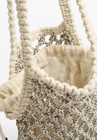 Neulepussi, jossa on vaalea crème-väri mustilla yksityiskohdilla, teksturoitu verkkomuotoilu ja pehmeä sisävuori. Mukana on letitetyt kahvat kantamista varten.