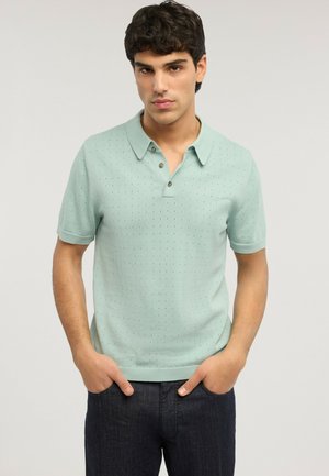 Giovane uomo che indossa una polo verde chiaro a maniche corte con piccole perforazioni, in piedi con le mani nelle tasche di un paio di jeans scuri.