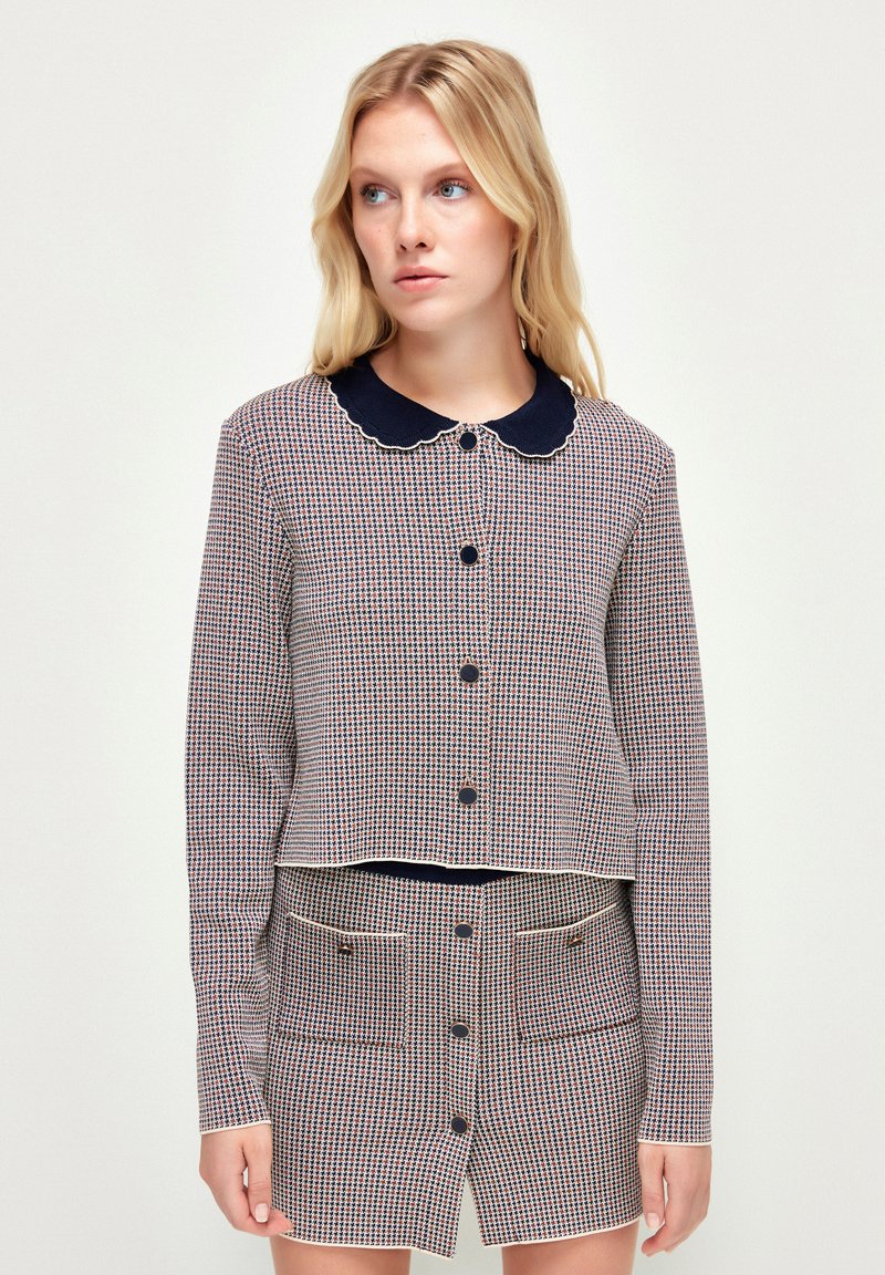 Stickad kort cardigan i ett rött och marinblått rutmönster med vågformig marinblå krage och knappar, i kombination med en matchande kjol som har fickor.