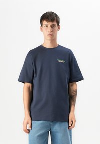 En ung mann iført en marineblå T-skjorte med teksten "London Fields Superstore" og lyseblå jeans, står mot en hvit bakgrunn.