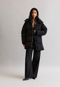 Schwarze Steppjacke mit gestepptem Design, hohem Kragen und Druckknöpfen. Trägt eine dunkle Outfit-Kombination, bestehend aus weiten Hosen und spitzen Schuhen.