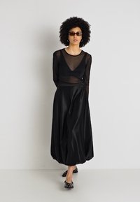 Fekete hálós, hosszú ujjú felső, amelyet egy könnyű fekete selyemszoknyával párosítottak. Az összeállítás mély dekoltázzsal rendelkezik és kényelmes, laza szabású.