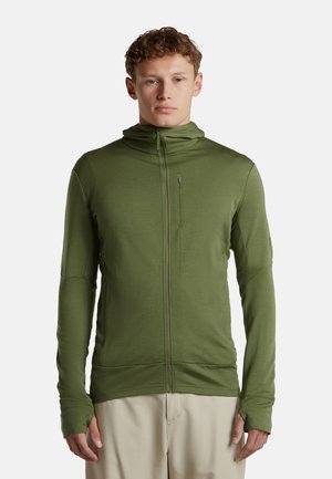 Joven con chaqueta verde oliva con cremallera y agujeros para el pulgar y pantalones beige, posando sobre un fondo liso.