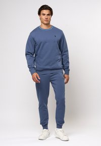 Modra dukserica in ujemajoče se trenirke iz mehkega materiala. Dukserica ima okroglo izreza s turkiznim robom; trenirke imajo stranske črte.