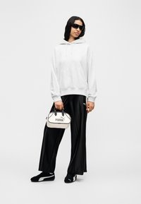 Γυναίκα που φοράει λευκή μπλούζα με κουκούλα, μαύρο παντελόνι με φαρδιά πόδια, μαύρα αθλητικά Puma, γυαλιά ηλίου με μεγάλο μέγεθος, κρατώντας λευκή τσάντα Puma, στέκεται σε λευκό φόντο.