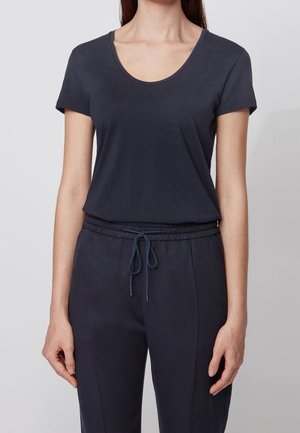 Kobieta ubrana w ciemnogranatowy t-shirt z krótkim rękawem i dekoltem w serek oraz pasujące spodnie ze sznurkiem, stojąca na tle jednolitego jasnego tła.