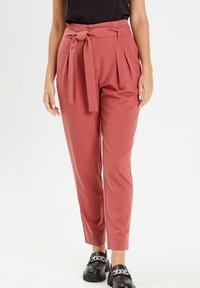 Pantalons en tissu rose doux, taille haute avec une ceinture à nouer, plis à l'avant, jambes fuselées et finition lisse. Chaussures noires avec détails en chaîne.