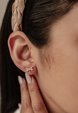 Gold dragonfly stud earring with clear gemstone accents, featuring a detailed wing design. Model's hand is gently touching the ear.