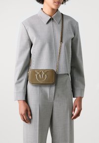 Pravougaona maslinasto zelena crossbody torba s reljefnim cvjetnim logotipom, zlatnom lancem za nošenje i kontrastnim šavovima. Model nosi košulju na kockice.