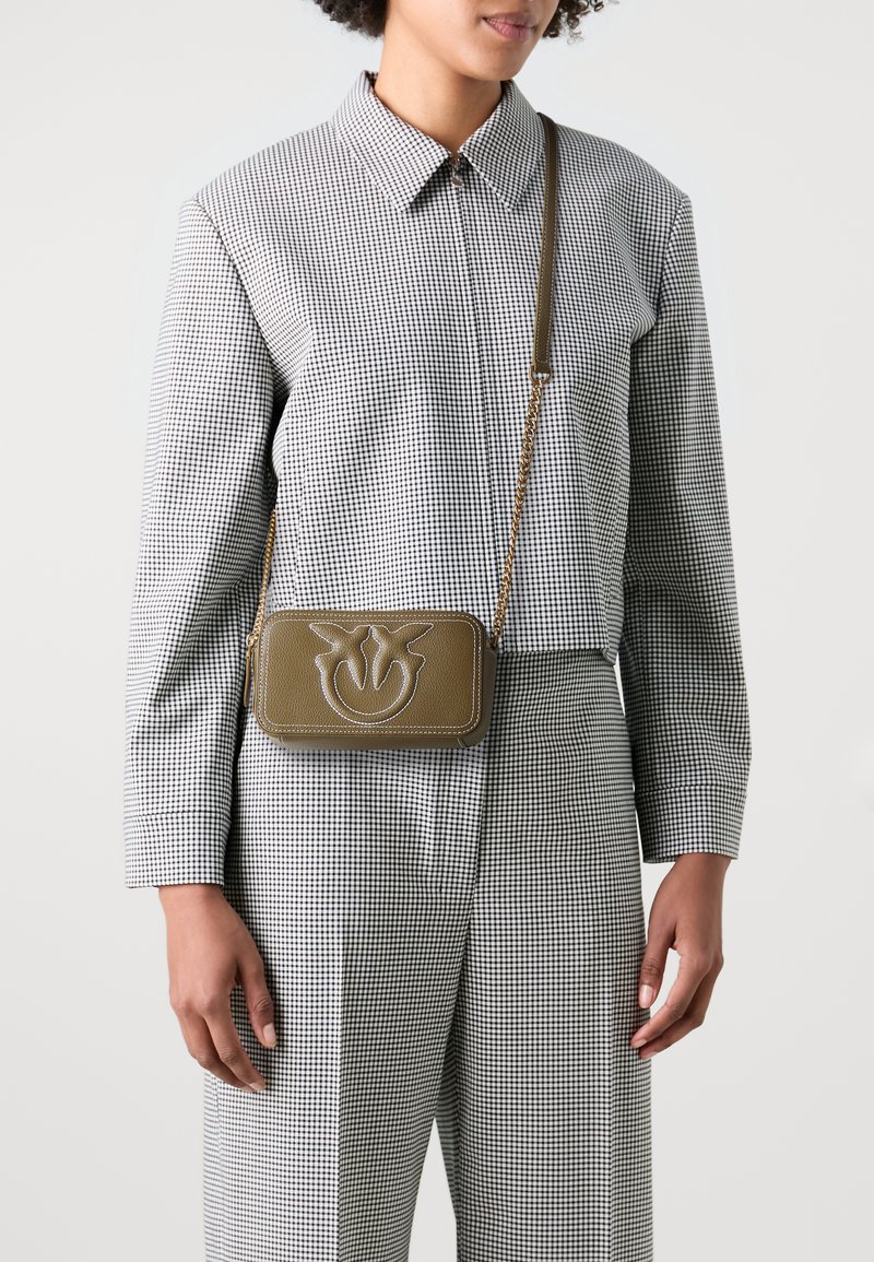 Pravougaona maslinasto zelena crossbody torba s reljefnim cvjetnim logotipom, zlatnom lancem za nošenje i kontrastnim šavovima. Model nosi košulju na kockice.